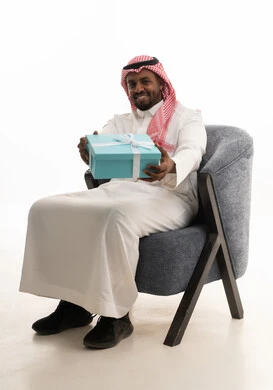 تقديم الهدايا بالاعياد والمناسبات الخاصة، التعبير عن الحب والتقدير والاحترام،بورتريه لرجل عربي خليجي سعودي يرتدي الثوب التقليدي والشماغ  يجلس على كرسي مريح يحمل بيديه صندوق هدية, ايماءات السرور والسعادة، مفهوم الشكر والامتنان، خلفية بيضاء