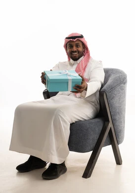 مفهوم الشكر والامتنان، تقديم الهدايا بالاعياد والمناسبات الخاصة، التعبير عن الحب والتقدير والاحترام،بورتريه لرجل عربي خليجي سعودي يرتدي الثوب التقليدي والشماغ  يجلس على كرسي مريح يحمل بيديه صندوق هدية, ايماءات السرور والسعادة، خلفية بيضاء