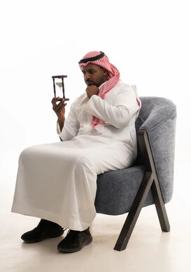 إدراك قيمة اللحظات الثمينة في الحياة، التخطيط لتحقيق الأهداف في المستقبل، انتظار شيء ما بفارغ الصبر، بورتريه لرجل عربي خليجي سعودي يرتدي الثوب التقليدي والشماغ يجلس على كرسي ويمسك الساعة الرملية، مراقبة مرور الوقت بايماءات السعادة والسرور، خلفية بيضاء