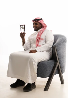 مراقبة مرور الوقت بايماءات السعادة والسرور، إدراك قيمة اللحظات الثمينة في الحياة، التخطيط لتحقيق الأهداف في المستقبل، انتظار شيء ما بفارغ الصبر، بورتريه لرجل عربي خليجي سعودي يرتدي الثوب التقليدي والشماغ يجلس على كرسي ويمسك الساعة الرملية، خلفية بيضاء