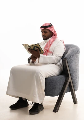 التقرب الى رب العالمين ، قراءة وحفظ القرآن الكريم،بورتريه لرجل عربي خليجي سعودي يرتدي الثوب التقليدي والشماغ  يجلس على كرسي مريح يقوم بقراءة المصحف الشريف، تلاوة كتاب الله تعالى ,الاجتهاد و الخشوع في العباده,  خلفية بيضاء