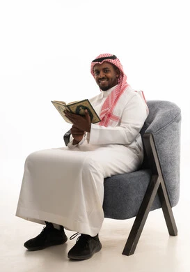 قراءة وحفظ القرآن الكريم،بورتريه لرجل عربي خليجي سعودي يرتدي الثوب التقليدي والشماغ  يجلس على كرسي مريح يقوم بقراءة المصحف الشريف، تلاوة كتاب الله تعالى ,الاجتهاد و الخشوع في العباده, التقرب الى رب العالمين ، خلفية بيضاء