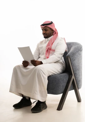 بورتريه لرجل عربي خليجي سعودي يرتدي الثوب التقليدي والشماغ يجلس على كرسي ينظر الى الاوراق البيضاء, التدقيق في قراءة الملاحظات، متابعة ومراجعة الخطط والمشاريع المكتبية، اعداد التقارير الخاصة بالعمل، خلفية بيضاء