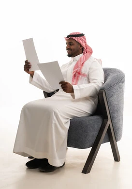 اعداد التقارير الخاصة بالعمل، متابعة ومراجعة الخطط والمشاريع المكتبية، بورتريه لرجل عربي خليجي سعودي يرتدي الثوب التقليدي والشماغ يجلس على كرسي ينظر الى الاوراق البيضاء, التدقيق في قراءة الملاحظات، خلفية بيضاء