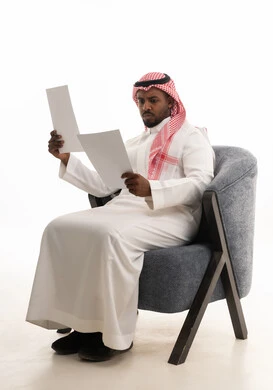 Monitoring and reviewing office plans and projects, preparing work-related reports, a portrait of a Saudi Arabian Gulf man wearing traditional attire and a ghutrah sitting on a chair looking at white papers, proofreading notes, white background.