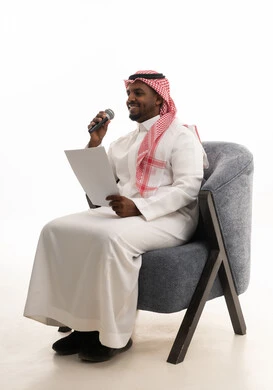 A portrait of a Saudi Arabian Gulf man wearing traditional attire and a shemagh sitting on a chair looking at white papers holding a microphone in his hand, checking notes, practicing singing talent, preparing work reports, following up and reviewing office plans and projects, with a white background.