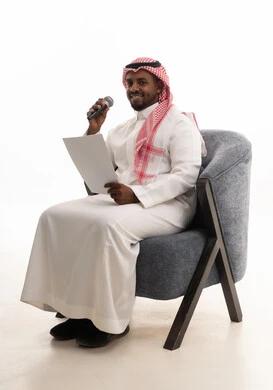 Presenting paragraphs using the microphone, preparing work-related reports, following up and reviewing office plans and projects, a portrait of a Saudi Gulf Arab man wearing traditional attire and a ghutrah sitting on a chair looking at white papers holding a microphone in his hand, proofreading notes, white background.