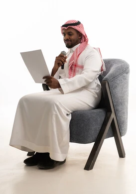 ممارسة موهبه الغناء، اعداد التقارير الخاصة بالعمل، متابعة ومراجعة الخطط والمشاريع المكتبية، بورتريه لرجل عربي خليجي سعودي يرتدي الثوب التقليدي والشماغ يجلس على كرسي ينظر الى الاوراق البيضاء يمسك بيده المايك, التدقيق في قراءة الملاحظات، خلفية بيضاء