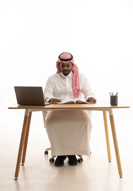 اجتماع عمل عن بعد، حب المطالعة والقراءة ،تمضية أوقات الفراغ بالتعلم والتثقف الذاتي, إيماءات الانتباه والتركيز،دمج التقنيات الحديثة فى الحياة اليومية ، إجراء بث مباشر مع الأهل والأصدقاء، بورتريه لرجل عربي خليجي سعودي يرتدي الثوب التقليدي والشماغ يجلس على مكتب يستخدم الحاسوب المحمول ، خلفية بيضاء