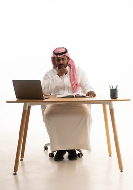 Integrating modern technologies into daily life, conducting live broadcasts with family and friends, remote work meetings, a love for reading and literature, spending free time learning and self-education, gestures of attention and focus, a portrait of an Arab Gulf Saudi man wearing traditional attire and a ghutrah sitting at a desk using a laptop, white background.