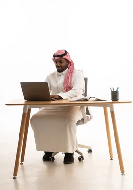 Spending free time on learning and self-education, a love for reading and literature, gestures of attention and focus, integrating modern technologies into daily life, conducting live broadcasts with family and friends, remote work meetings, a portrait of an Arab Gulf Saudi man wearing traditional attire and a ghutrah sitting at a desk using a laptop, white background.