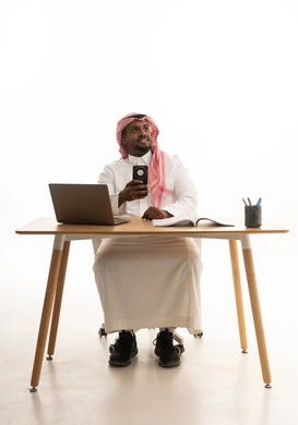 النظر جانباً بايماءات السعادة ، دمج التقنيات الحديثة فى الحياة اليومية ، استخدام الهاتف المحمول ، إجراء بث مباشر مع الأهل والأصدقاء، اجتماع عمل عن بعد، بورتريه لرجل عربي خليجي سعودي يرتدي الثوب التقليدي والشماغ يمسك الجوال بيده يستخدم الحاسوب المحمول ، خلفية بيضاء