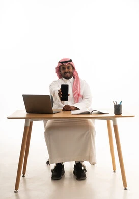 A portrait of a Saudi Arabian Gulf man wearing traditional attire and a ghutrah, holding a mobile phone in one hand and using a laptop, integrating modern technologies into daily life, using a mobile phone, conducting a live broadcast with family and friends, remote work meeting, white background.