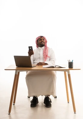 Conducting a live broadcast with family and friends, a remote work meeting, integrating modern technologies into daily life, using a mobile phone, a portrait of a Saudi Arabian Gulf man wearing traditional attire and a ghutrah holding a mobile phone in his hand while using a laptop, white background.