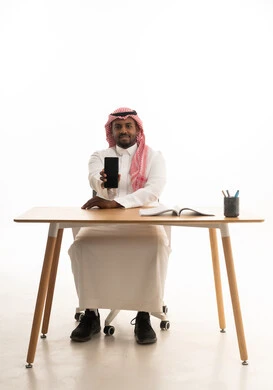 عرض شاشة سوداء فارغة ، دمج التقنيات الحديثة فى الحياة اليومية ، استخدام الهاتف المحمول ، إجراء بث مباشر مع الأهل والأصدقاء، تصفح مواقع التواصل الاجتماعي ، بورتريه لرجل عربي خليجي سعودي يرتدي الثوب التقليدي والشماغ يمسك الجوال بيده  وامامه كتاب ، خلفية بيضاء