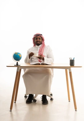 إيماءات السرور والسعادة ، الاستمتاع بالمشروبات الساخنة ، حب المطالعة والقراءة ،تمضية أوقات الفراغ بالتعلم والتثقف الذاتي,بورتريه لرجل عربي خليجي سعودي يرتدي الثوب التقليدي والشماغ يجلس على مكتب يقوم بقراءه كتاب وبيده فنجان من القهوه  ، خلفية بيضاء