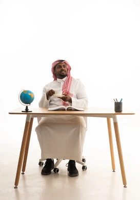 Spending free time learning and self-educating, a love for reading and literature, gestures of attention and focus, enjoying hot beverages, a portrait of an Arab Gulf Saudi man wearing traditional attire and a ghutrah sitting at a desk reading a book with a cup of coffee in his hand, white background.