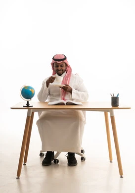 حب المطالعة والقراءة ،تمضية أوقات الفراغ بالتعلم والتثقف الذاتي, إيماءات الانتباه والتركيز، الاستمتاع بالمشروبات الساخنة ، بورتريه لرجل عربي خليجي سعودي يرتدي الثوب التقليدي والشماغ يجلس على مكتب يقوم بقراءه كتاب وبيده فنجان من القهوه  ، خلفية بيضاء