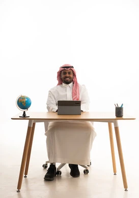 اجتماع عمل عن بعد، بورتريه لرجل عربي خليجي سعودي يرتدي الثوب التقليدي والشماغ يجلس على مكتب يمسك جهاز التابلت ينظر بإيماءات السعادة، إجراء مكالمات الفيديو مع الأهل والأصدقاء،خلفية بيضاء