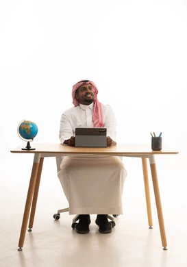 إجراء مكالمات الفيديو مع الأهل والأصدقاء، اجتماع عمل عن بعد، بورتريه لرجل عربي خليجي سعودي يرتدي الثوب التقليدي والشماغ يجلس على مكتب يمسك جهاز التابلت ينظر بإيماءات السعادة، خلفية بيضاء