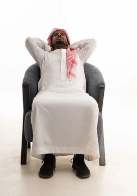 The need for sleep and the feeling of tiredness and lethargy, expressions of contemplation and thought, a portrait of an Arab Gulf Saudi man wearing traditional attire and a ghutrah sitting on a comfortable chair with his hands behind his head in gestures of comfort and relaxation, white background.