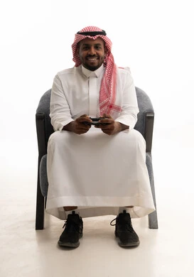 The use of modern electronic entertainment devices and technologies, interaction and immersion with video games, spending enjoyable and entertaining time, a portrait of a Saudi Arabian Gulf man wearing traditional attire and a headscarf holding a game controller, with a white background.