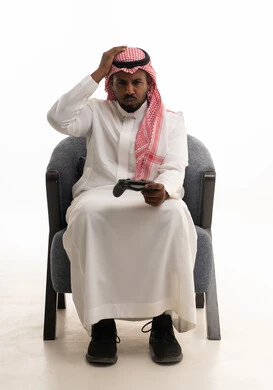 وضع اليد على الرأس بتعابير الهزيمة ،التفاعل والاندماج مع ألعاب الفيديو، قضاء وقت ممتع ومسلي، استخدام اجهزة وتقنيات الكترونية ترفيهيه حديثة,  بورتريه لرجل عربي خليجي سعودي يرتدي الثوب التقليدي والشماغ يمسك بيده عصا التحكم، خلفية بيضاء .