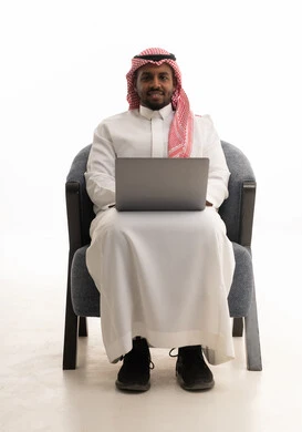 اجتماع عمل عن بعد، إجراء مكالمات الفيديو مع الأهل والأصدقاء،بورتريه لرجل عربي خليجي سعودي يرتدي الثوب التقليدي والشماغ يمسك جهاز الحاسوب المحمول وينظر إليه بإيماءات السعادة، خلفية بيضاء