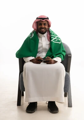 أنديه رياضية مختلفة، الحماس والتفاعل مع المنتخب، إيماءات الشغف والتشجيع للفريق، بورتريه لرجل عربي خليجي سعودي مبتسم يرتدي الشماغ والثوب التقليدي والعلم السعودي يجلس على كرسي مريح يشاهد مباراة كرة القدم ، خلفية بيضاء .