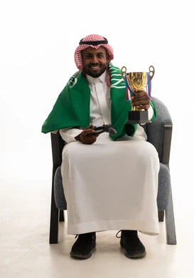إيماءات الشغف والتشجيع للفريق، الحماس والتفاعل مع المنتخب، مشاهدة ومتابعة مباراة كرة القدم، بورتريه لرجل عربي خليجي سعودي مبتسم يرتدي الشماغ والثوب التقليدي والعلم السعودي يجلس على كرسي مريح يحمل كأس المنتخب، أنديه رياضية مختلفة، خلفية بيضاء .