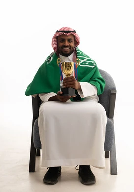 الحماس والتفاعل مع المنتخب، إيماءات الشغف والتشجيع للفريق، مشاهدة ومتابعة مباراة كرة القدم، بورتريه لرجل عربي خليجي سعودي مبتسم يرتدي الشماغ والثوب التقليدي والعلم السعودي يجلس على كرسي مريح يحمل كأس المنتخب، أنديه رياضية مختلفة، خلفية بيضاء .