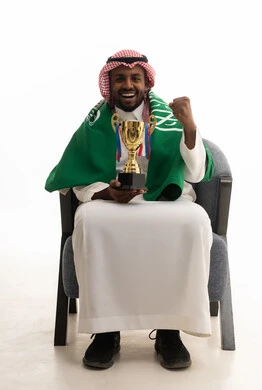 Raising a fist with gestures of enthusiasm and interaction with the national team, gestures of passion and encouragement for the team, watching and following a football match, a portrait of a smiling Arab Gulf Saudi man wearing a ghutrah and traditional thobe, holding the Saudi flag while sitting on a comfortable chair with the national team's cup, different sports clubs, white background.