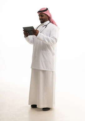 A portrait of a smiling Saudi Gulf doctor wearing a ghutrah and traditional attire along with a white coat, looking at the camera while providing services to patients with great efficiency and using a tablet, utilizing modern technologies in work, the concept of medicine and healthcare, with a white background.