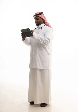 The concept of medicine and healthcare, the use of modern technologies in work, a portrait of a smiling Saudi Gulf doctor wearing a ghutrah and traditional attire along with a white coat, looking at the camera, providing services to patients with efficiency and using a tablet, with a white background.