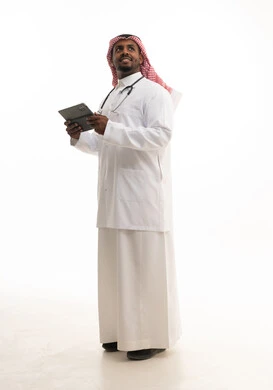 التواصل مع المرضي عن بعد ، مفهوم الطب والرعاية الصحية ، بورتريه لطبيبب سعودي خليجي مبتسم يرتدي الشماغ والزي التقليدي والمعطف الابيض ينظر للكاميرا يقدم خدماته للمرضى بكل كفاءه ويستخدم الجهاز اللوحي،  خلفية بيضاء