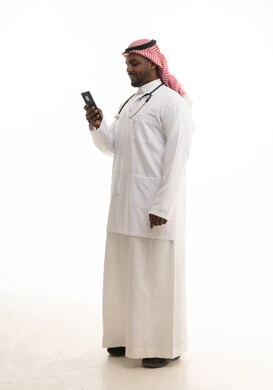 مفهوم الطب والرعاية الصحية ، استخدام التقنيات الحديثة فى العمل ، بورتريه لطبيبب سعودي خليجي مبتسم يرتدي الشماغ والزي التقليدي والمعطف الابيض ينظر للكاميرا يقدم خدماته للمرضى بكل كفاءه ويستخدم الهاتف المحمول،  خلفية بيضاء