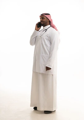Integrating modern technology into work, the concept of medicine and healthcare, a portrait of a smiling Saudi Gulf doctor wearing a ghutrah and traditional attire along with a white coat, looking at the camera, providing services to patients with efficiency and using a mobile phone, with a white background.