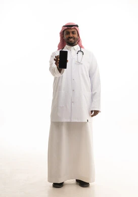 The use of modern technologies in work, the concept of medicine and healthcare, a portrait of a smiling Saudi Gulf doctor wearing a ghutrah and traditional attire along with a white coat, looking at the camera, providing services to patients with efficiency and using a mobile phone, white background.