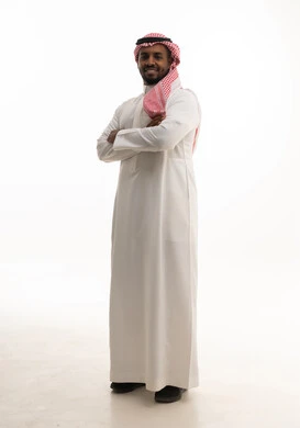 الوقوف بايدي متقاطعة ، مفهوم الرجولة والقوة والثقة بالنفس، بورتريه لرجل عربي خليجي سعودي يرتدي الشماغ والزي التقليدي ينظر الى الكاميرا بإيماءات السرور والسعادة ، الاهتمام بالأناقة والمظهر الخارجي ، خلفية بيضاء