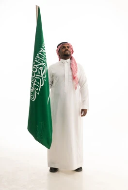 الاحتفال بذكرى اليوم الوطني السعودي 8 ربيع الأول, يوم العلم 11مارس،الفخر و الاعتزاز بالانتماء للمملكة, بورتريه لرجل عربي سعودي خليجي مبتسم يرتدي الشماغ والثوب التقليدي يحمل بيده علم السعودية , خلفية بيضاء