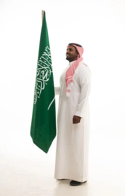 يوم العلم 11مارس، الاحتفال بذكرى اليوم الوطني السعودي 8 ربيع الأول, الفخر و الاعتزاز بالانتماء للمملكة, بورتريه لرجل عربي سعودي خليجي مبتسم يرتدي الشماغ والثوب التقليدي يحمل بيده علم السعودية , خلفية بيضاء