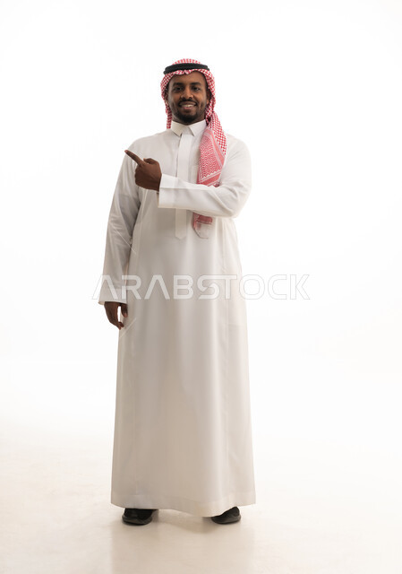 حركات تدل على هذا المكان (هنا), الدلالة على موقع معين,تحديد مكان عنصر محدد, بورتريه لرجل عربي خليجي سعودي مبتسم يرتدي الشماغ والثوب التقليدي يشير بإصبع السبابة إلى شيء ما, الجسم بالطول الكامل, خلفية بيضاء