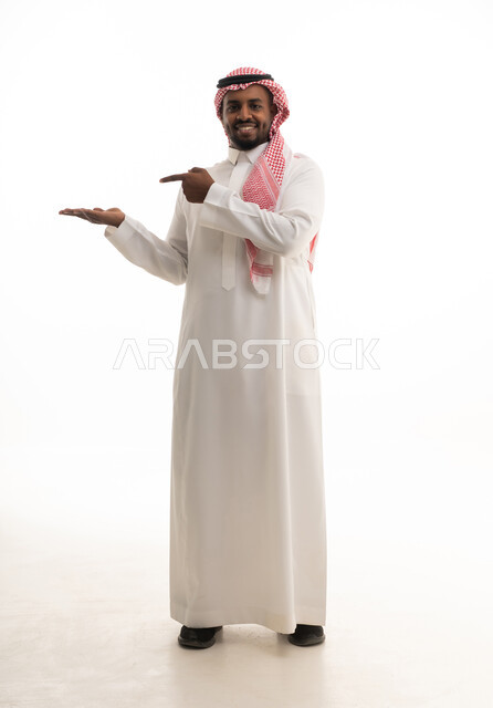 تعابير الفرح والسرور والاشاره الى شئ ما،بورتريه لرجل عربي خليجي سعودي مبتسم يرتدي الشماغ والثوب التقليدي يشير بإصبع السبابة ليده الأخرى، الامساك بعنصر ما بإيماءات الاعجاب،  خلفية بيضاء