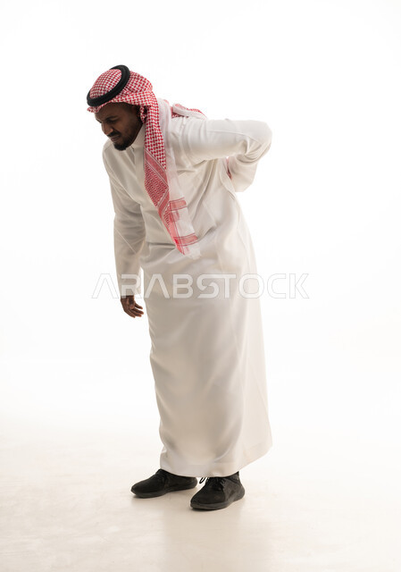 آلام العمود الفقري والظهر، بورتريه لرجل عربي خليجي سعودي مبتسم يرتدي الشماغ والثوب التقليدي يضع يده على ظهره بتعابير الانحناء، إيماءات الوجع والألم، التعب والإرهاق والإحساس بالمرض، الحاجة إلى العلاج وزيارة الطبيب، صورة الجسم بالكامل، خلفية بيضاء