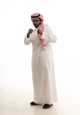 إتقان فنون الملاكمة، تعزيز صحة العظام والمفاصل، ممارسة رياضة الدفاع عن النفس، زيادة قدرة العضلات على التحمل، بورتريه لرجل عربي خليجي سعودي يرتدي الثوب التقليدي والشماغ  يرفع قبضات يديه بإيماءات القوة، الطول الكامل للجسم، خلفية بيضاء