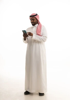 Conducting a live broadcast with family and friends, browsing social media, integrating modern technologies into daily life, using a mobile phone, a portrait of a Saudi Arabian Gulf man wearing traditional attire and a ghutrah holding a mobile phone in his hand, white background.
