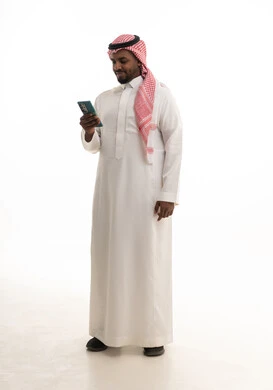 Browsing social media, a portrait of a Saudi Arabian Gulf man wearing traditional attire and a ghutrah holding a mobile phone in his hand, integrating modern technologies into daily life, using a mobile phone, conducting a live broadcast with family and friends, white background.