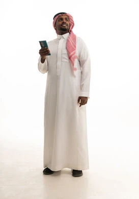 دمج التقنيات الحديثة فى الحياة اليومية ، استخدام الهاتف المحمول ، إجراء بث مباشر مع الأهل والأصدقاء، تصفح مواقع التواصل الاجتماعي ، بورتريه لرجل عربي خليجي سعودي يرتدي الثوب التقليدي والشماغ يمسك الجوال بيده ، خلفية بيضاء
