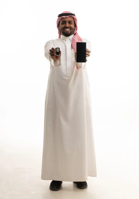 عرض شاشة سوداء فارغة عبر  الجوال ، النظر إلى الكاميرا بإيماءات السعادة و السرور, تعابير الفرح لامتلاك سيارة جديدة, بورتريه لرجل عربي خليجي سعودي مبتسم يرتدي الثوب التقليدي و الشماغ يمسك بيده مفتاح التحكم عن بعد, صورة للجسم بالطول الكامل, خلفية بيضاء