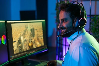 استخدام برامج المونتاج المتقدمة، صورة من الجانب لمونتير عربي خليجي سعودي يرتدي الثوب الابيض يضع سماعة الراس يعمل في غرفة المونتاج يستخدم الحاسب المحمول لقص وترتيب المشاهد، مفهوم صناعة المحتوى المرئي والإبداع الفني